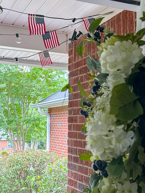 Coastal Americana front porch decorated with DIY American flag string light garland on ceiling and white hydrangea wreath on blue front door