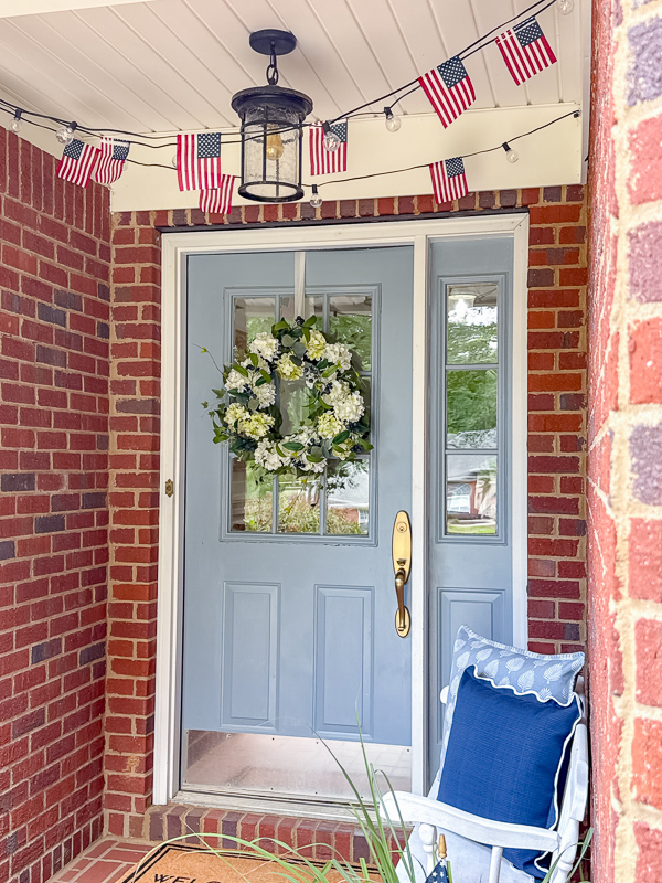 Coastal Americana front porch decorated with DIY American flag string light garland on ceiling and white hydrangea wreath on blue front door