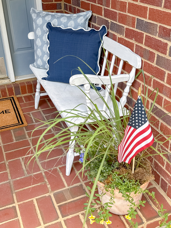 Red brick porch with white bench and navy blue pillow and light blue pattern pillow with lemon grass plant beside it with flowers and an stick American flag in the planter.