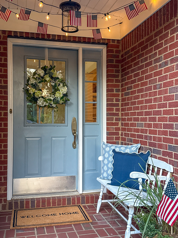 Porch at dusk with American flag string light along the ceiling, blue door with white and green hydrangea wreath, bench with blue pillows and container garden with flag in it.