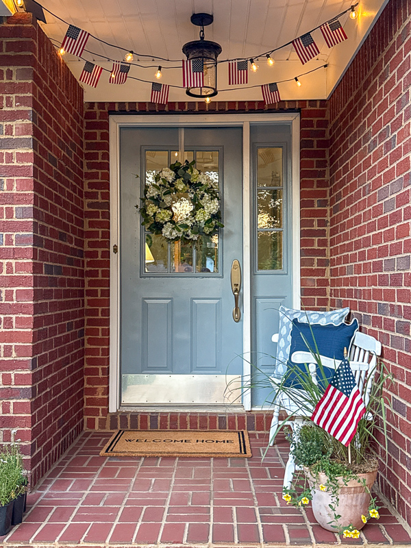 View at dusk of a patriotic porch with American flag string light garland across the ceiling, white bench with blue pillows, container garden with an American flag in it a classic hydrangia wreath on a blue door.