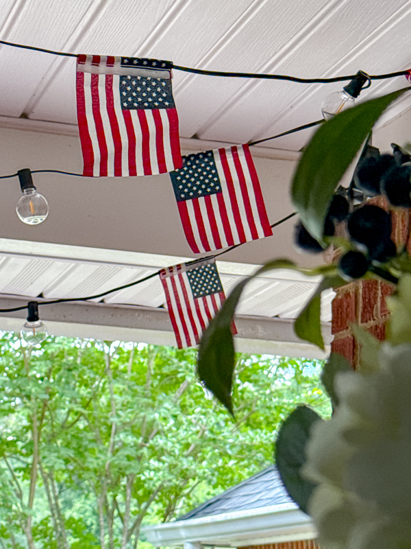 Coastal Americana front porch decorated with DIY American flag string light garland on ceiling and white hydrangea wreath on blue front door