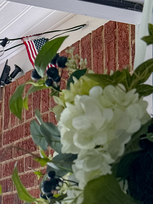 DIY American flag string light garland hanging on covered porch ceiling with white hydrangea wreath on front door. Showing a hook to hang the garland.