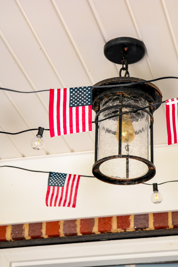 DIY American flag solar string light garland draped around black porch lantern on white beadboard ceiling