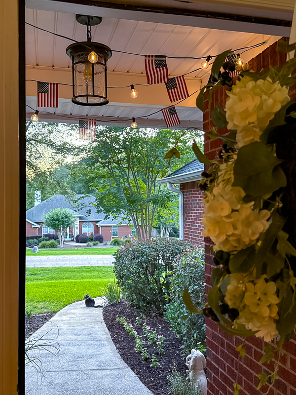 DIY American flag string light garland glowing at dusk on covered front porch with hydrangea wreath and patriotic decor
