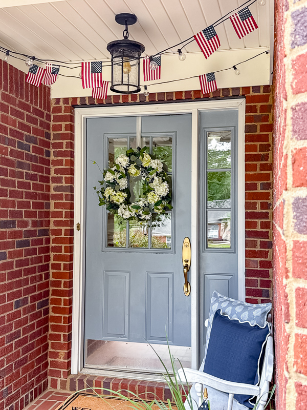 Small porch during the day with American flag string light along the ceiling, blue door with white and green hydrangea wreath, bench with blue pillows.