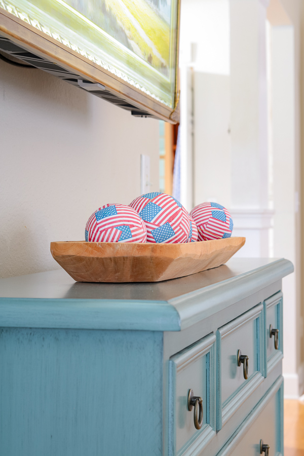 Wooden dough bowl filled with mini American flag balls. Sittong on a blue piece of furniture