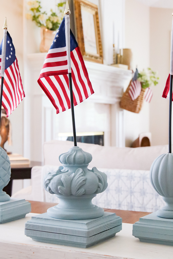 DIY patriotic finial flag stand painted in Dixie Belle Savannah Mist chalk paint with a small Betsy Ross flag displayed on a white coffee table with a cozy living room and fireplace mantel in the background