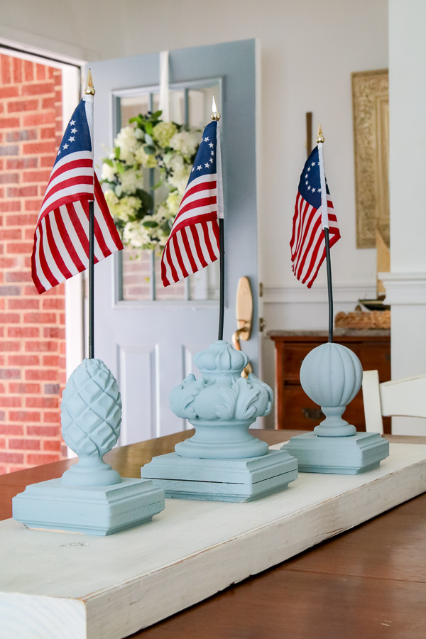 Three DIY patriotic finial flag stands painted in Dixie Belle Savannah Mist chalk paint grouped together as a 4th of July table centerpiece in a cozy French country cottage foyer with a blue door in the background