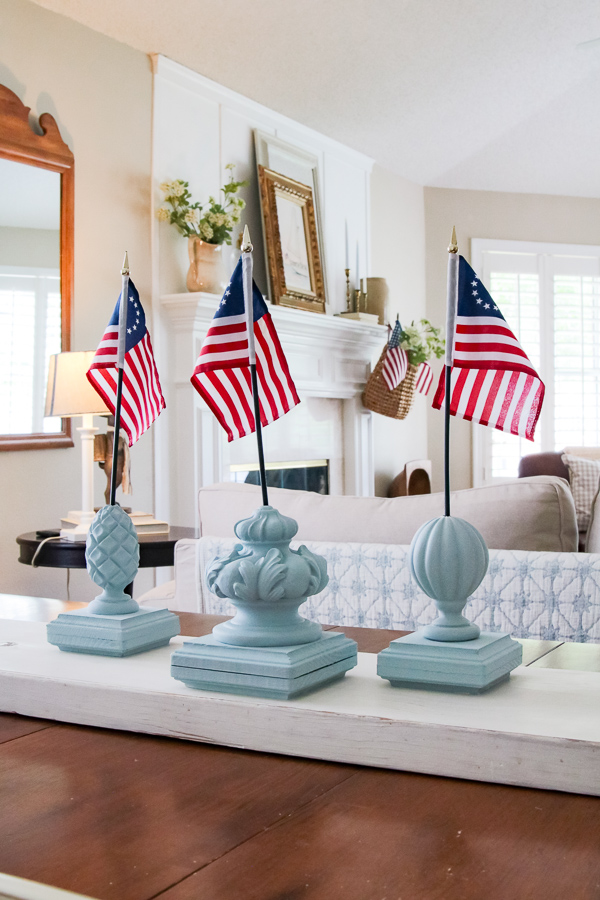 Three DIY patriotic finial flag stands painted in Dixie Belle Savannah Mist chalk paint grouped together as a 4th of July table centerpiece in a cozy French country cottage living room with a white fireplace mantel in the background