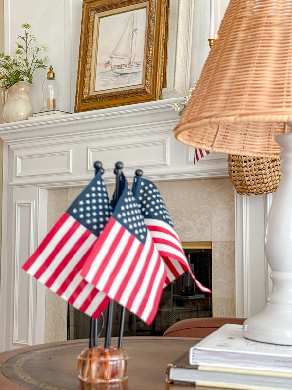 Watercolor sailboat print in gold frame hung on white board and batten wall with American flags in brass candlesticks on stacked vintage books