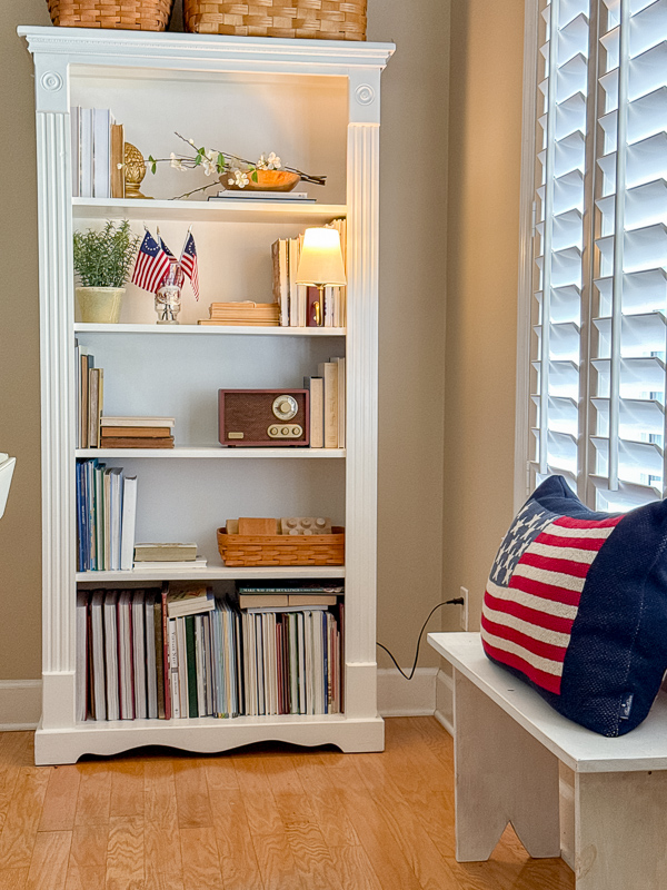 White bookcase styled with vintage books, small American flags, brass accents and woven baskets for year-round Americana decor