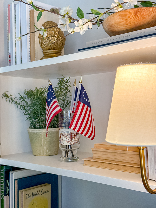 Vintage bookcase shelf styled with Betsy Ross flags in a vintage 200th Anniversary glass, rosemary plant, stacked books and a small lamp for Ralph Lauren Americana decor