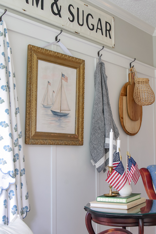 Watercolor sailboat print in gold frame hung on white board and batten wall with American flags in brass candlesticks on stacked vintage books