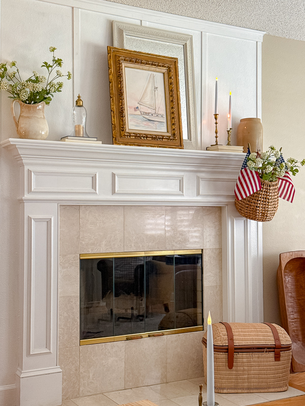Year-round Americana decor styled on a white fireplace mantel with a watercolor sailboat print in a gold frame, brass candlesticks, a woven basket with American flags and white flowers