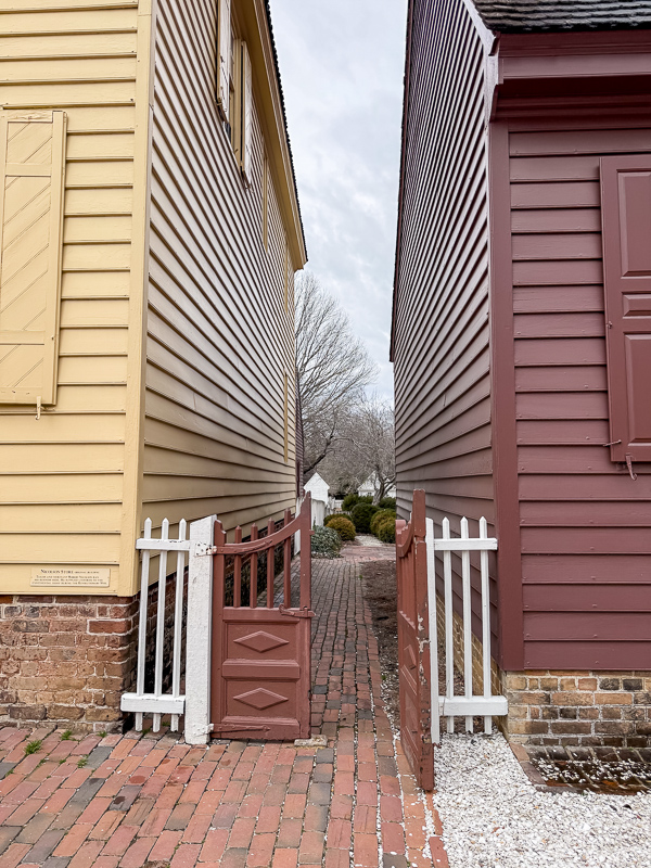 Alley at Williamsburg with white picket fence on side of a red gate that has a picket on top and diamond pattern on bottom.