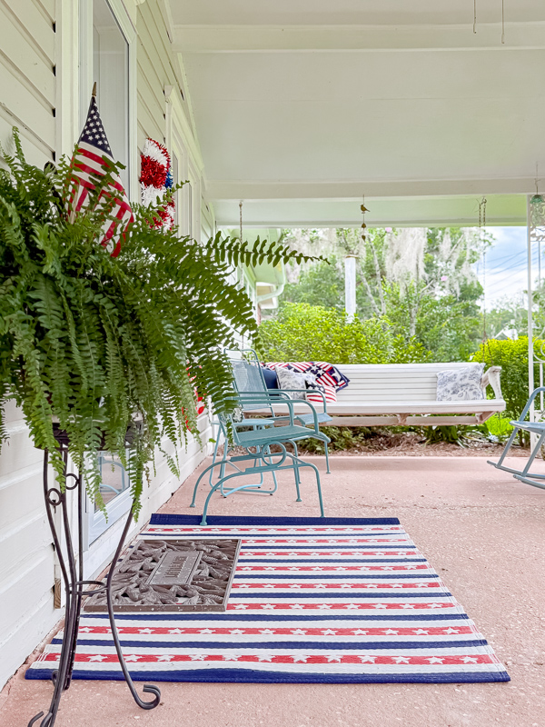 Large porch patriotic decor ideas using red, white and blue rug at the front door laytered with a iron doormat on top.