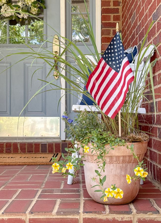 Patriotic porch decor ideas. Terracotta planter with plants and a stick flag in front of a white bench and a blue door with a hydrangea wreath.