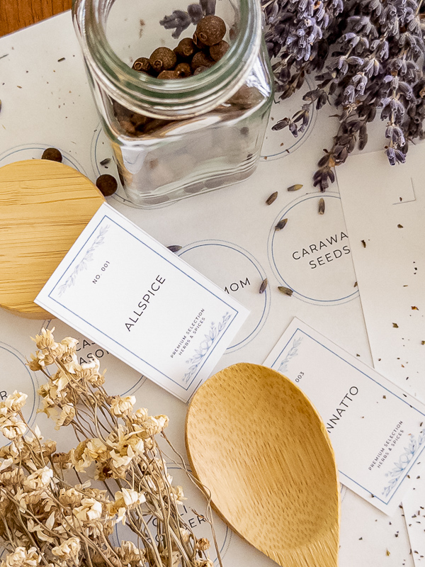 Close-up of vintage blue and white spice label printables including allspice and annatto, styled with an open jar of allspice, dried lavender, and a wooden spoon