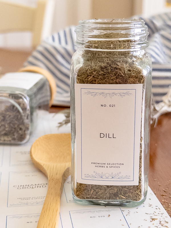 Glass jar of dill filled to the top with a vintage printable spice label, styled with a wooden spoon and blue striped linen