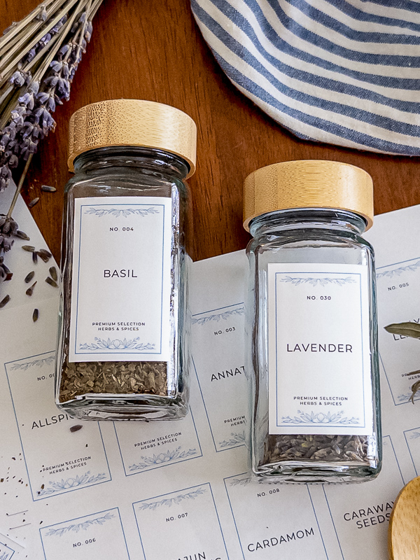 Basil and lavender jars with vintage printable spice labels resting on a sheet of uncut labels, styled with dried lavender, blue striped linen, and a wooden spoon