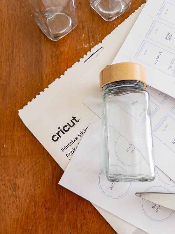 Cricut printable sticker paper with vintage spice labels, an empty glass spice jar, and a pencil ready for tracing and cutting