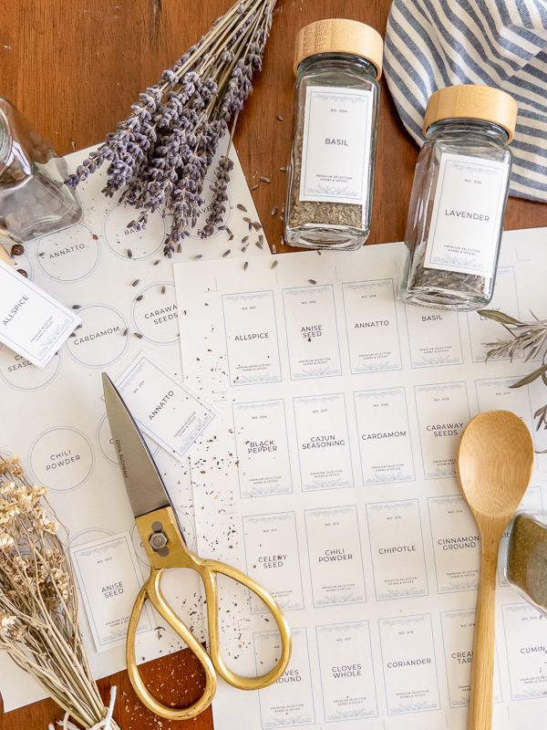 Printed vintage spice label sheets being cut with gold scissors and applied to glass spice jars, styled with dried lavender and a wooden spoon