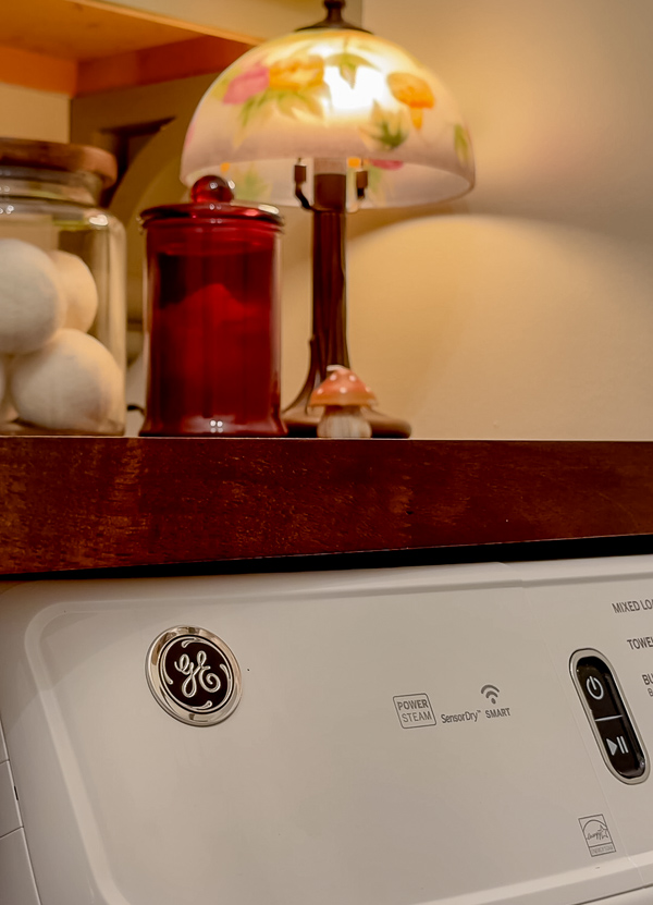 GE washer, vintage lamp, candle and large jar with wool balls on a butcher block countertop. Whimsical laundry room.