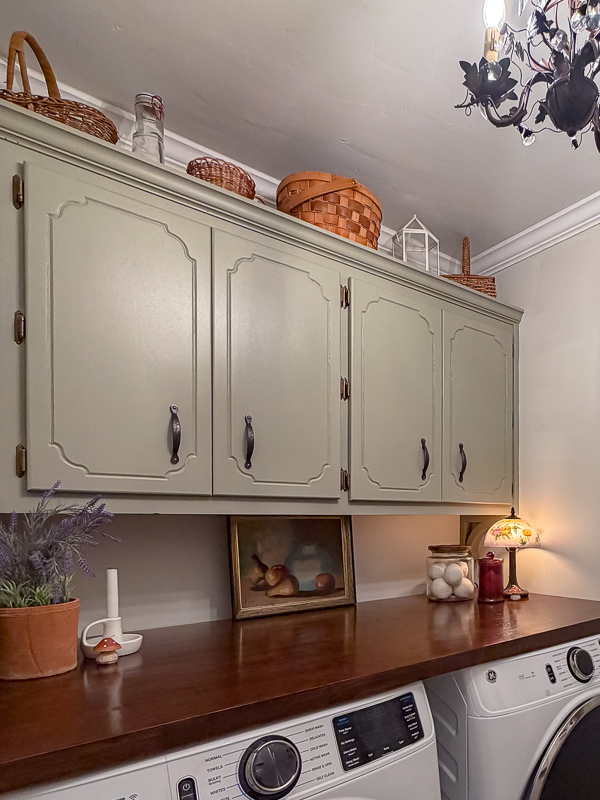 Cozy and whimsical cottage garden theme laundry room makeover with painted green cabinets, bronze ivy hardware, art on butcher block countertop stained in walnut, ge front load washer and dryer and baskets on top of cabinet.