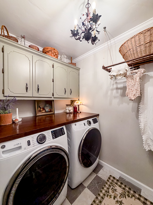 Cozy and whimsical cottage garden theme laundry room makeover with painted green cabinets, bronze ivy hardware, art on butcher block countertop stained in walnut, ge front load washer and dryer and baskets on top of cabinet and homemade Shiela Maid.