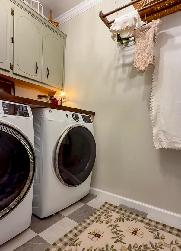 Whimsical cottage garden laundry room. Green cabinets, baskets, Shiela Maid, vintage lamp, Lowe's lavender plant, GE front load washer and dryer and glass jar, butcher block countertop and bee motif rug.