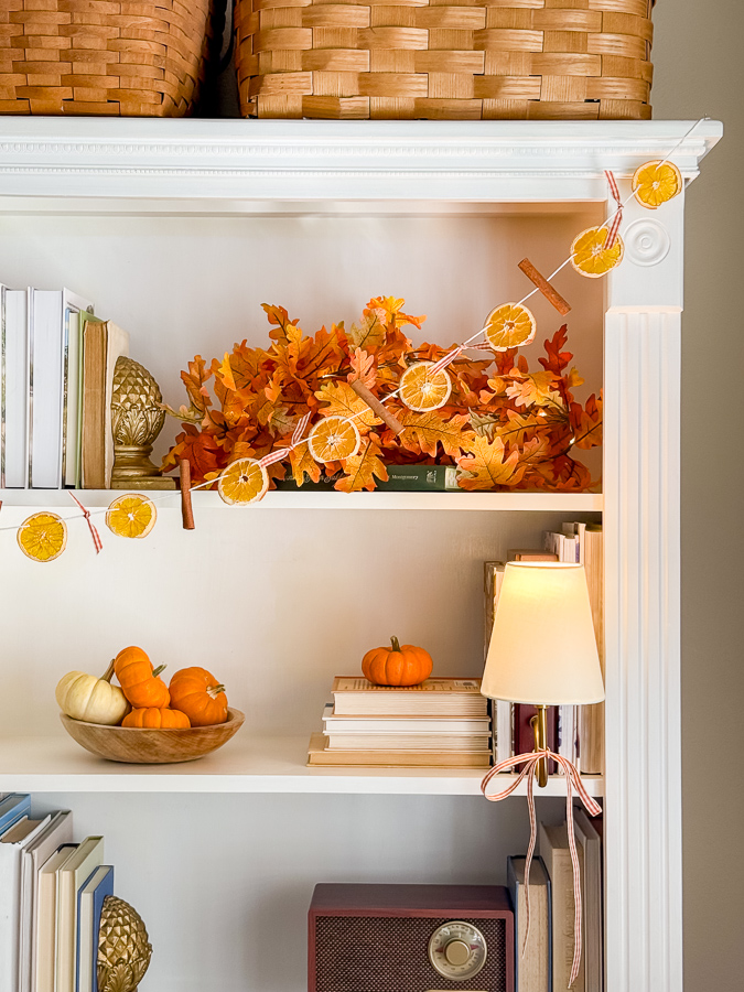 Dried orange fall garland with cinnamon sticks and gingham bows swagged across a white bookcase with fall leaves and pumpkins