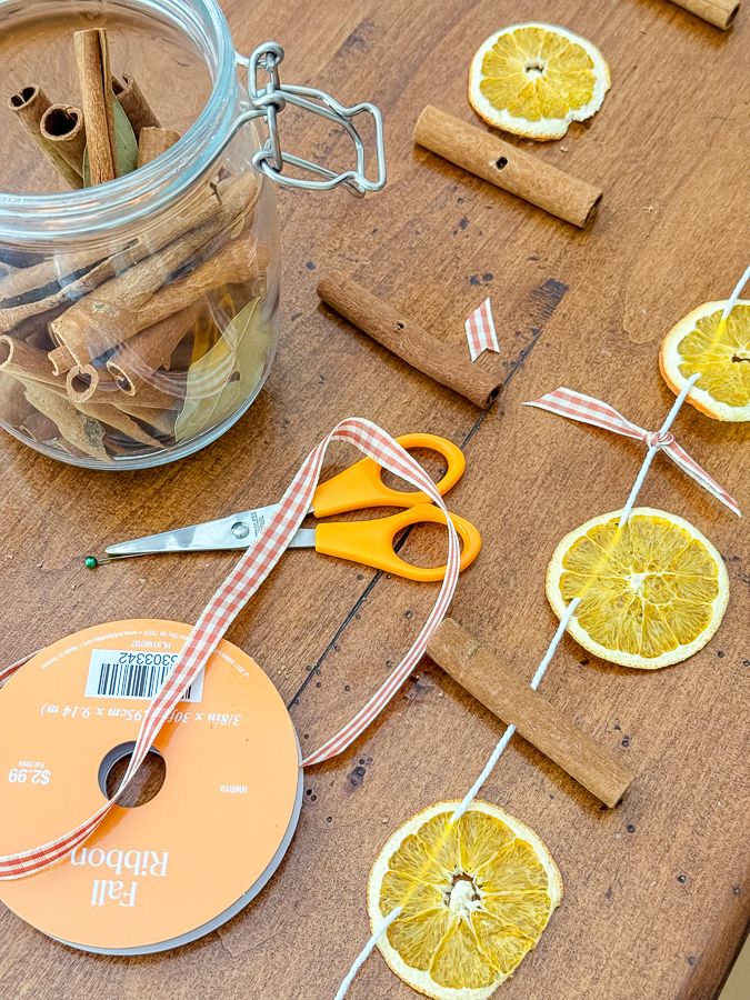 Garland supplies on a wooden table: dried orange slices, cinnamon sticks with drilled holes, gingham ribbon, and scissors