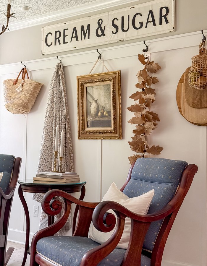 Sitting room corner with a vintage Cream and Sugar sign, straw market basket, block-print tablecloth, gilt-framed pear print, and paper leaf garland on wall hooks