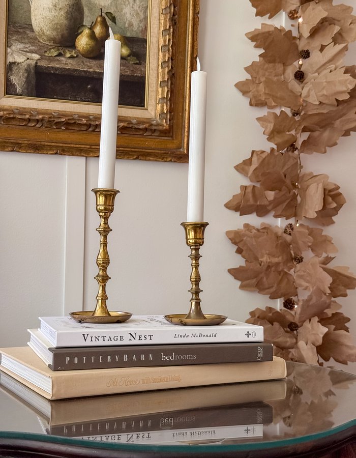 Gilt-framed vintage-style art print of an ironstone pitcher and pears, hanging above brass candlesticks and the book Vintage Nest