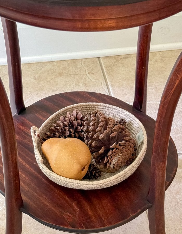 A small rope bowl holding a faux pear and pinecones, tucked on the side table beneath the brass candlesticks