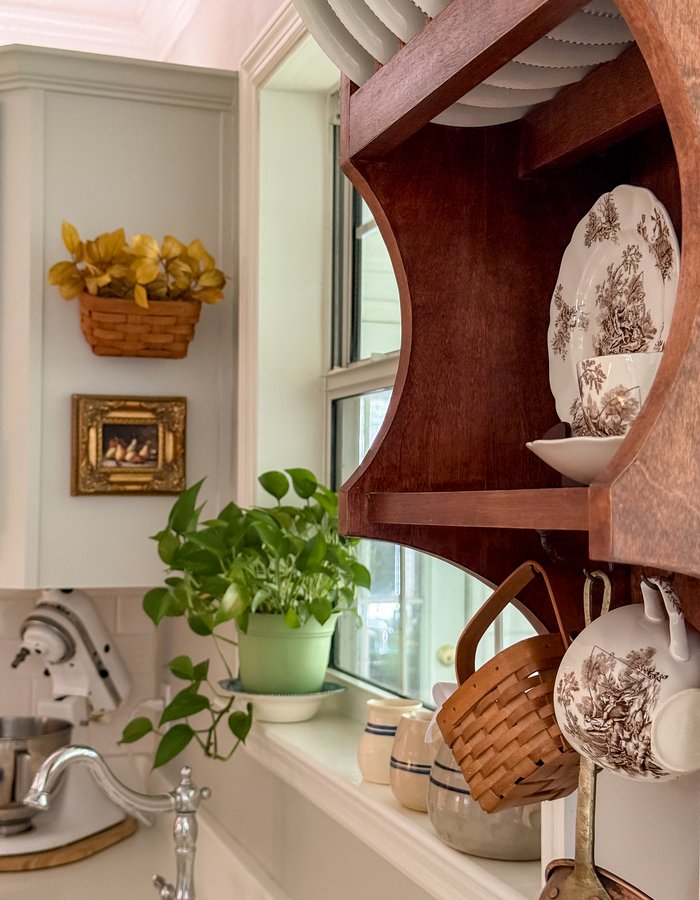 Wooden shelf above the kitchen sink styled with brown transferware plates, a toile teacup, and a hanging garlic basket by the window