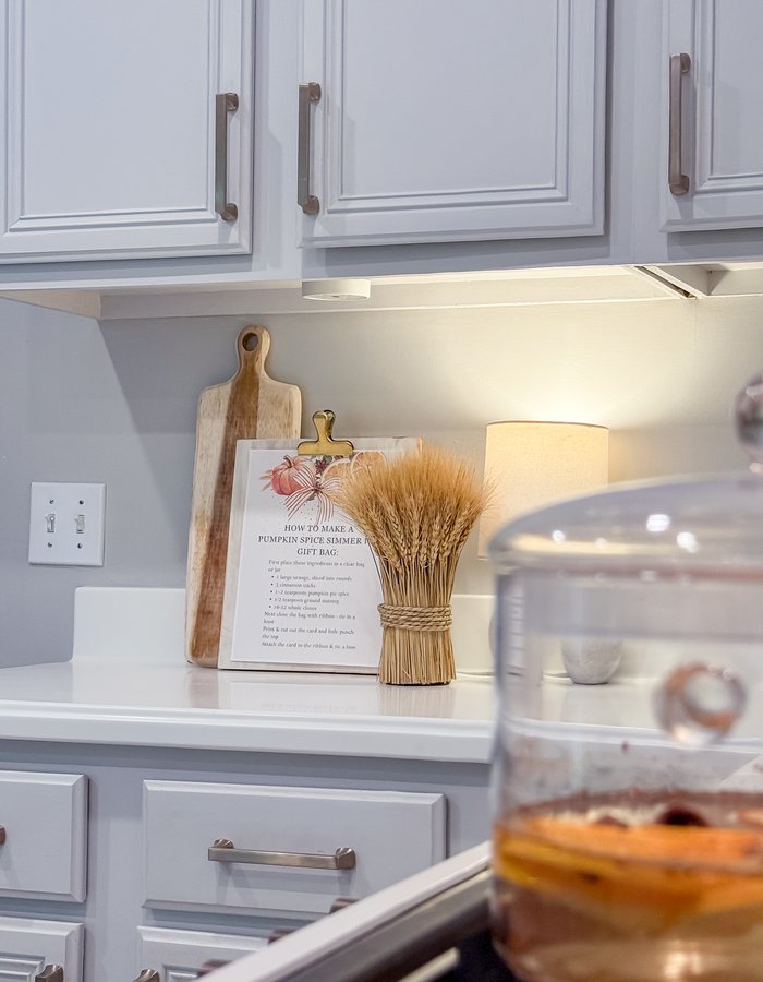 Wide view of the kitchen counter fall vignette with cutting boards, the wheat bundle, and the lamp glowing softly under the cabinets