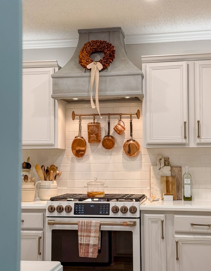 French country range hood decorated for fall with a rusty boxwood wreath, hanging copper pots, and a plaid towel over the oven handle
