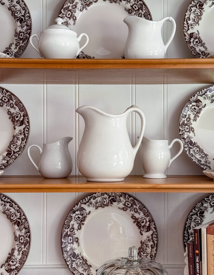 White ironstone pitchers in graduated sizes lined up on the hutch shelf with brown and white Spode plates behind them