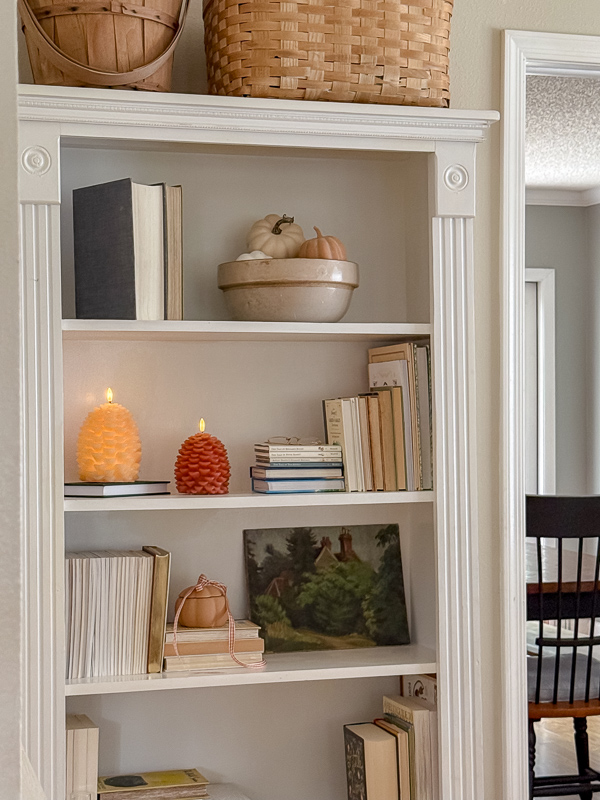 Bookcase styled with a bowl of white pumpkins, burnt orange and white pinecone candles, and stacked books for fall