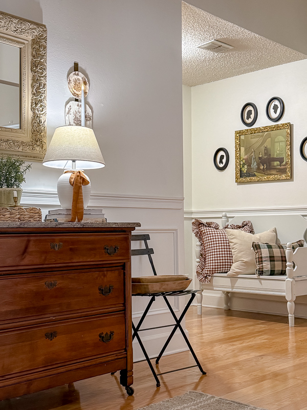 Fall entryway vignette with an antique wood dresser, table lamp, and a gallery wall of vintage oval frames flanking a gilt-framed painting