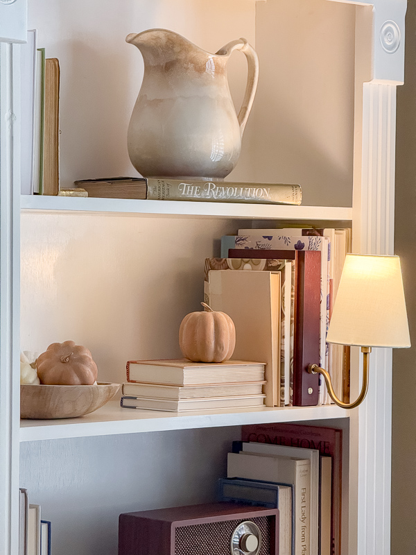 Fall bookcase styling with a stoneware pitcher, stacked books, and mini pumpkins in a green and brown fall decor palette