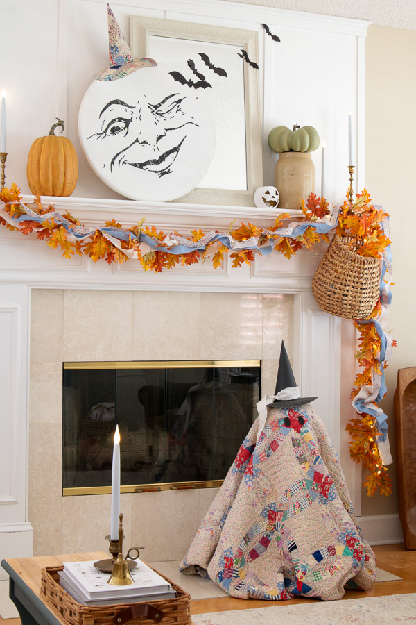 Cozy cottage Halloween decor mantel with a man in the moon face, lighted fall leaf garland, and a vintage quilt ghost by the fireplace