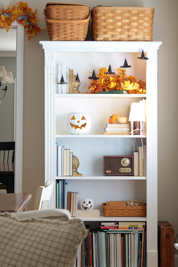 White bookcase decorated for Halloween with a mini witch hat garland, white jack-o'-lanterns, and baskets