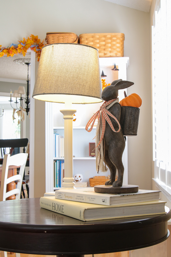 Bunny statue carrying a pumpkin in its basket with an orange gingham ribbon beside a lamp on an end table