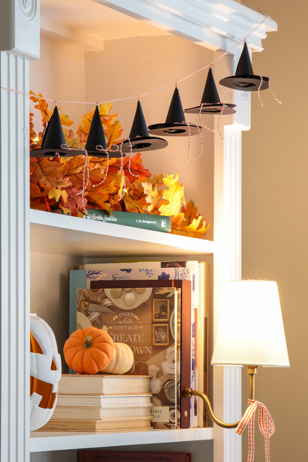 Mini paper witch hat garland on a bookcase shelf with lighted fall leaves