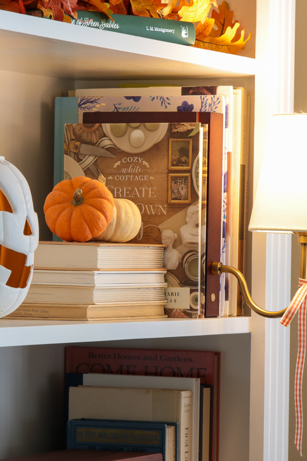 Real orange and white baby boo pumpkins on stacked vintage books in a bookcase beside a white jack-o'-lantern