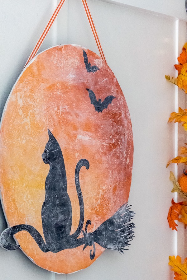 Black cat riding a broom in front of a glowing harvest moon paper door hanger.